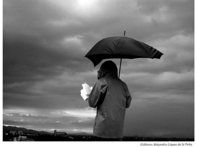 Black and white photograph of a person, back to the camera, holding a broken umbrella over their head as they read. The sky is cloudy and the city is visible in the distance.