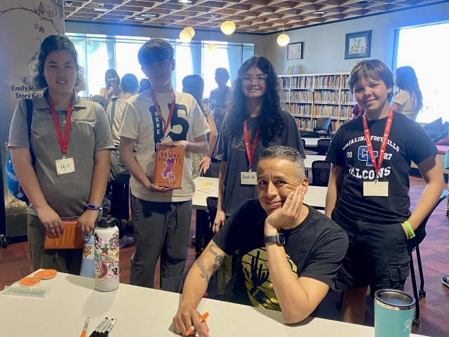 Four teens stand behind a smiling man who signs their books