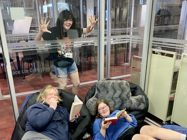 Two teens lounge as they read while another teen mock surprises them from the other side of a glass wall.