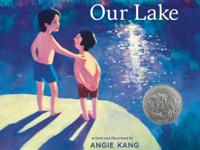 Two boys in swim trunks stand in front of a blue lake.