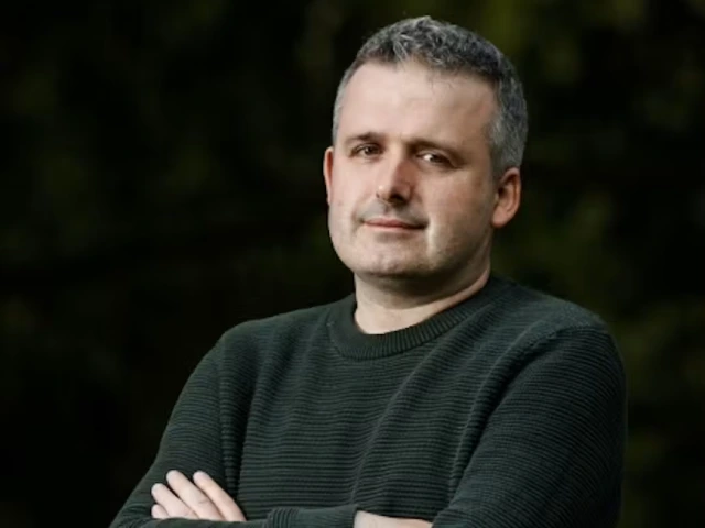 A white man in a grey-green sweater crosses his arms.