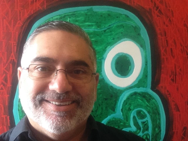 Head shot of Darryn Joseph. Behind him is one of his paintings using bold strokes in green and red.