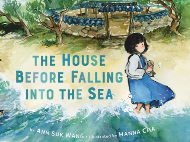 A young girl in a blue dress stands on a beach with her feet in the ocean. Behind her is the gate going to her small house.