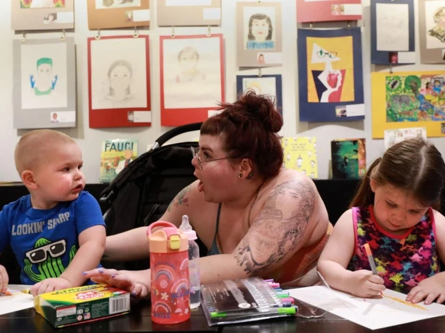An aunt enthusiastically engages her two children in creating art against a backdrop of children's self-portraits.