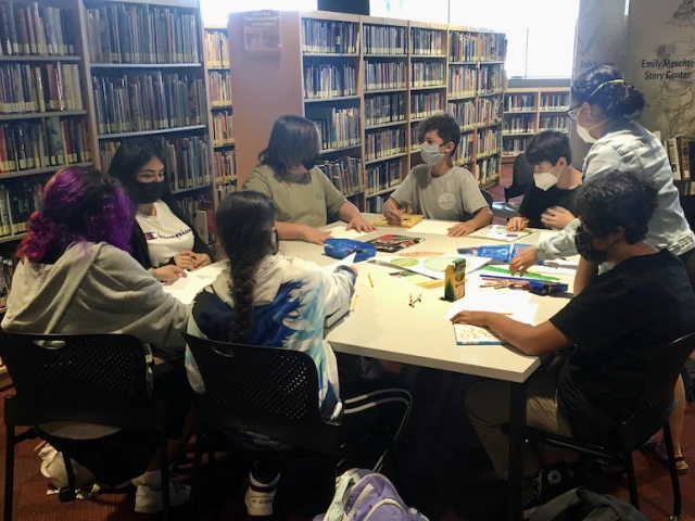 Younger teens sit around a large table creating heart and mind maps using paper, color pencils, crayons, markers and rulers.