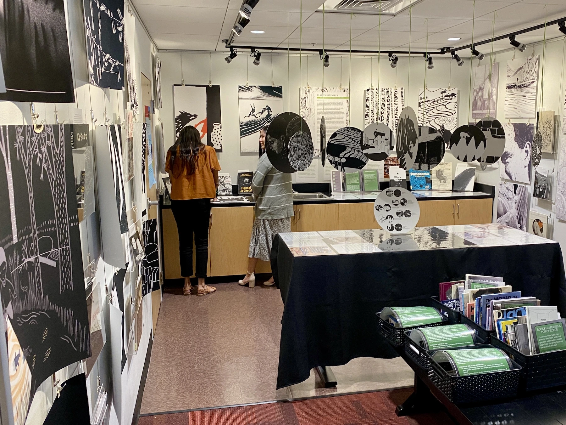 Two women study a puzzle in a corner of a room filled with black and white illustrations