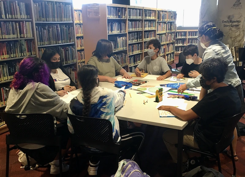 Younger teens sit around a large table creating heart and mind maps using paper, color pencils, crayons, markers and rulers.