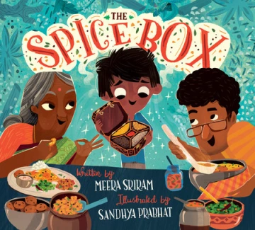 A young Indian boy holds open a box full of spices. Next to him are his dad and gradnmother are smiling at him. They all stand in front of a table full of food.