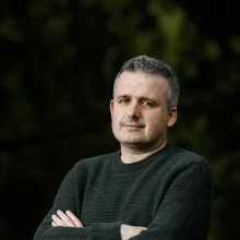 A white man in a grey-green sweater crosses his arms.