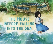 A young girl in a blue dress stands on a beach with her feet in the ocean. Behind her is the gate going to her small house.
