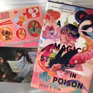 Photo of the novel A Magic Steeped in Poison next to an illustration of the characters and a sticker sheet inspired by the book.