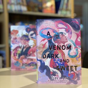 Photo of the novel A Venom Dark and Sweet, with book A Magic Steeped in Poison in the background in front of a bookshelf.