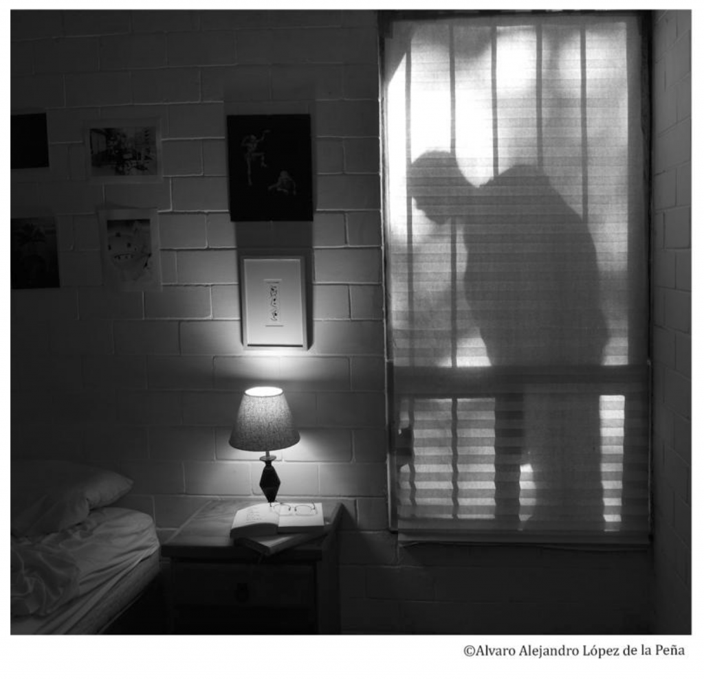 The shadow of a man in a window peers into an empty bedroom, a book open face up on a nightstand. The photograph is in black and white.