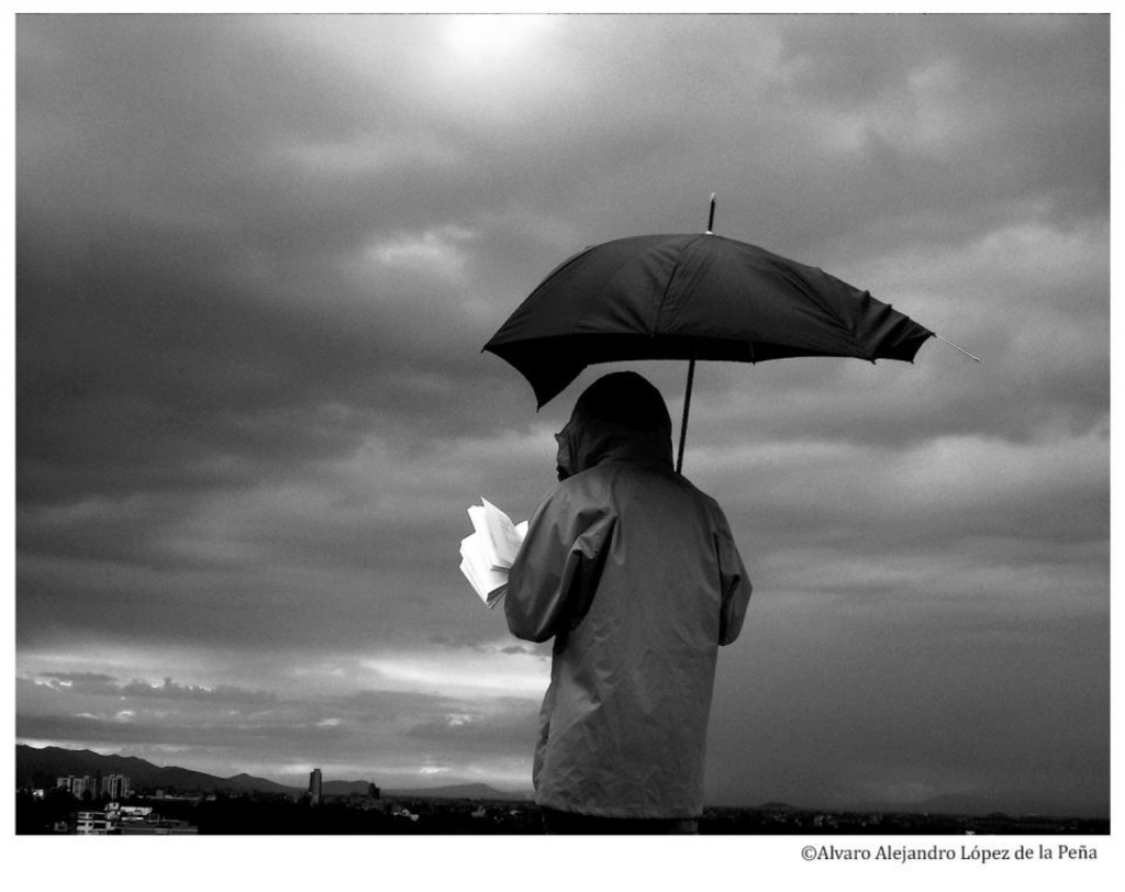 Black and white photograph of a person, back to the camera, holding a broken umbrella over their head as they read. The sky is cloudy and the city is visible in the distance.