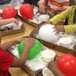 Image of a group of four young children rolling balloons in a tray of flour. In the back are two red balloons, while the children closer to the camera ar eusing green (left side) and white (right side).