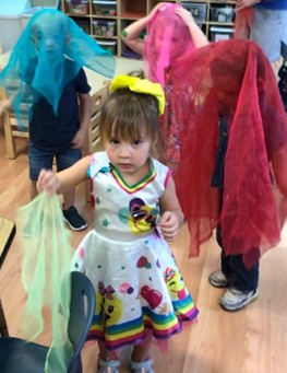 A girl in a rainbow dress holds out a scarf