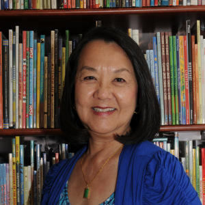 A smiling Mary J. Wong posed in front of a shelf with many picturebooks.