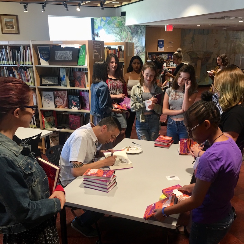 Matt Mendez signs copies of Barely Missing Everything at a table surrounded by teen readers