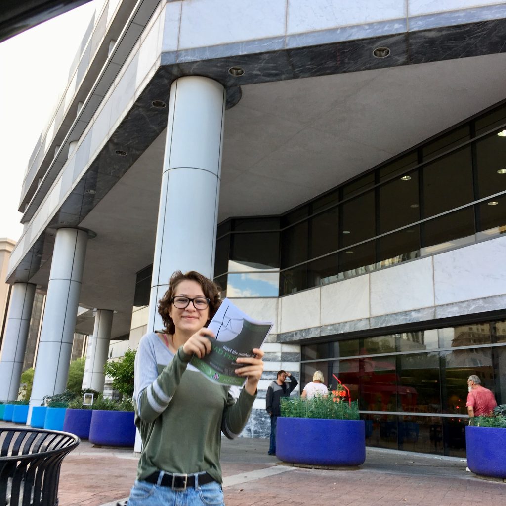 A Teen Reading Ambassador holds up book flyers in front of a public library.