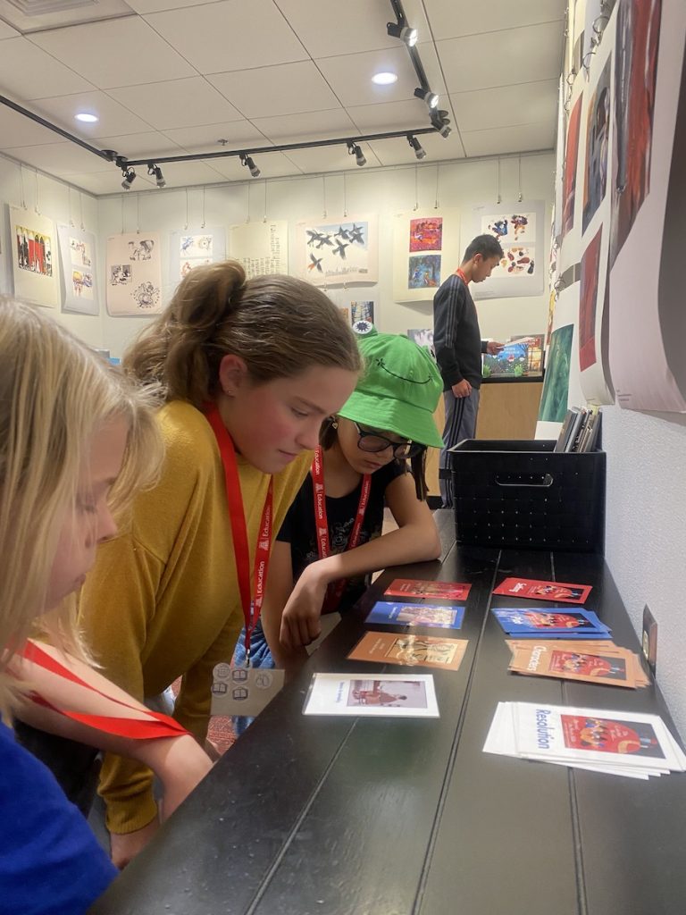 Three young people look at a set of cards marked character, setting, plot and resolution one young person in background looking at illustrations