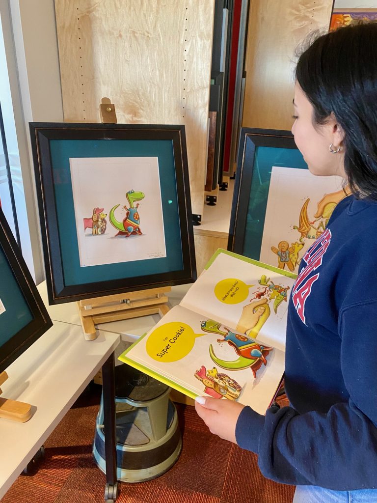 Young woman compares original illustrations of a superhero dinosaur cookie to a final published version.