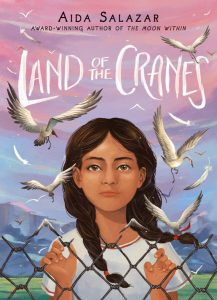 Cover of Land of the Cranes, depicting a young girl with two braided pigtails looking at the viewer with her fingers curled around the gaps in a chain link fence. Small cranes fly around her face and the background in a pink to light blue gradient sky and mountains.